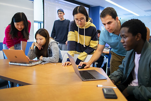 classroom with diverse group of students gathered around laptops